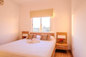 A hotel room with a large bed, wooden headboard, bedside tables, and a window with a view. - 2BR Sunrise Port View Apt Steps from Finikoudes (Larnaka)