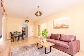 A modern living room with a sofa, a coffee table, and a dining area with chairs and a table. - 2BR Sunrise Port View Apt Steps from Finikoudes (Larnaka)