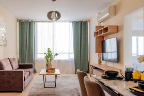 A modern living room with a sofa, a dining table, a TV, and a kitchen area. - 2BR Sunrise Port View Apt Steps from Finikoudes (Larnaka)