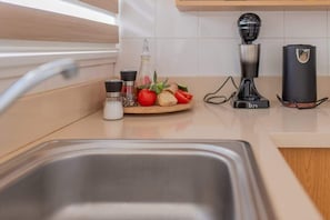 Eine Küche mit Spüle aus Edelstahl, einem Holztisch mit Tomaten und Kräutern, einer Kaffeemaschine und einer Flasche Olivenöl.