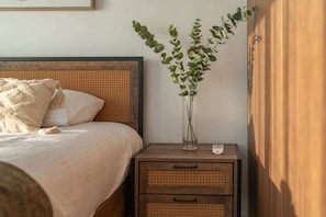Ein Bett mit Holz-Kopfstütze, ein Nachttisch mit einer Vase mit grünen Blättern und eine kleine Kommode.