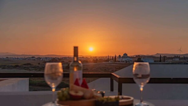 Ein Tisch mit zwei Weingläsern und einer Flasche, vor einer Sonnenuntergangslandschaft.