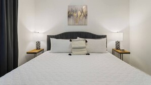 Una cama bien hecha con cabecera, dos mesitas de noche con lámparas, y un cuadro en la pared.
