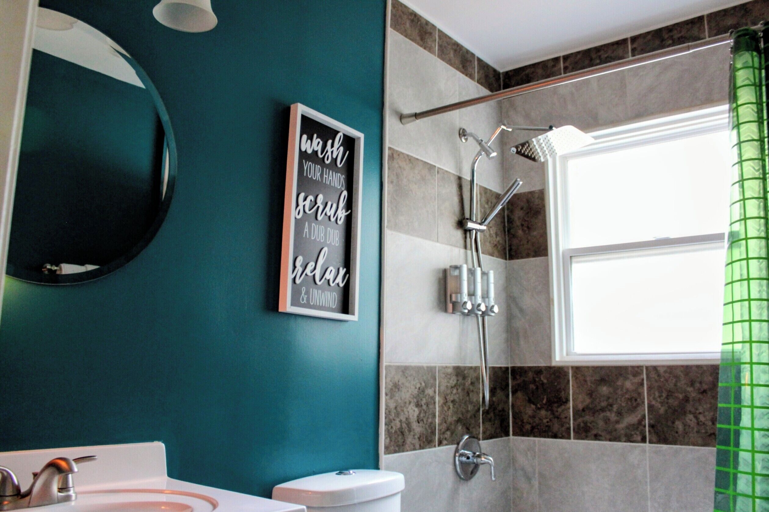 A bathroom with a teal wall, a framed sign, a shower, and a window with a green curtain.