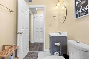 A bathroom with a toilet, sink, and mirror. There is a wooden shelf with a bottle and a towel rack. A door leads to another room. - The Queen City Hub 3 (Cincinnati)