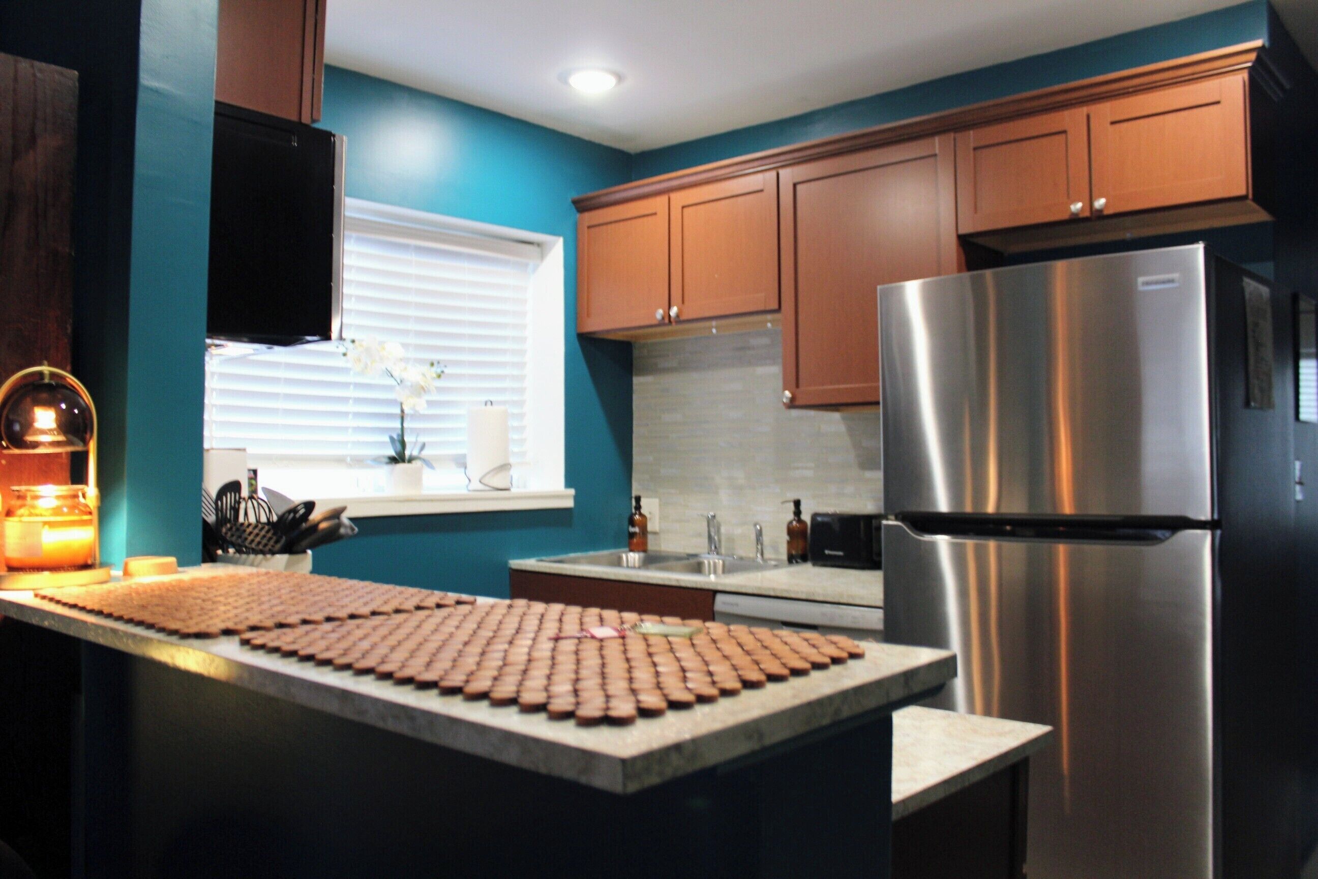 A kitchen with wooden cabinets, a stainless steel refrigerator, a window with blinds, and a countertop with a cutting board.