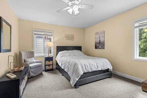 A bedroom with a bed, a chair, a nightstand, a lamp, a ceiling fan, a window, and a wall with a picture. - The Queen City Hub 3 (Cincinnati)