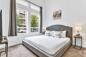 A bedroom with a large bed, two bedside lamps, a window with curtains, and a painting on the wall. - Quiet Getaway | Weekly & Monthly Discounts (Richardson)