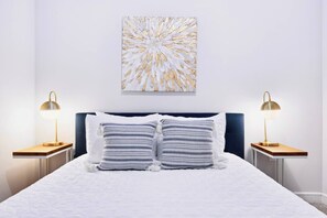Ein Bett mit weißer Bettwäsche, zwei Nachttischlampen und ein an der Wand hängendes abstraktes Gemälde.