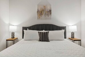 A bed with a quilted white cover, two bedside lamps, a black and white patterned pillow, and a painting above the bed. - Near Trails & Eats | Weekly & Monthly Specials (Richardson)