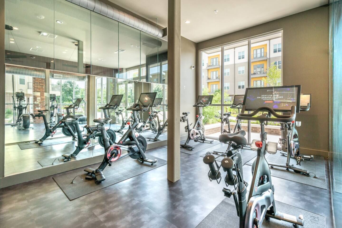 Un gimnasio con bicicletas estáticas, espejos y un ventanal con vistas a edificios.