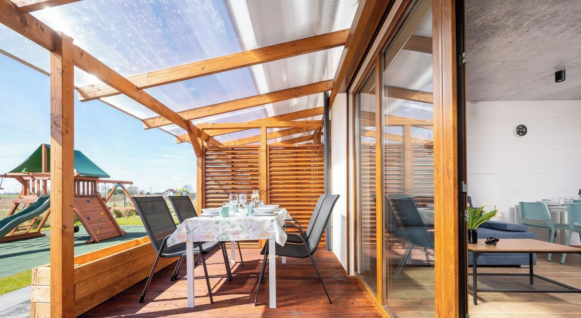 A covered patio with a table and chairs, a wooden play structure, and a glass door leading to an indoor area.