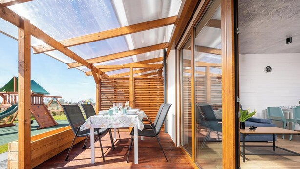 A covered patio with a table and chairs, a wooden play structure, and a glass door leading to an indoor area. - Pleasant 2-bedroom house with AC in charming Mielenko (Mielenko)