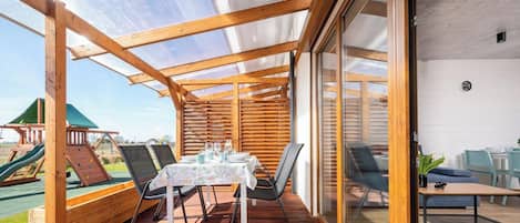 Un porche cubierto con mesa y sillas, un parque infantil de madera y una puerta de cristal que da a un espacio interior.