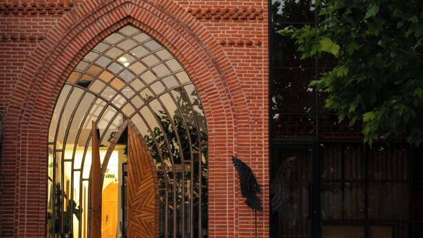 A brick building with a large arched entrance, a glass door, and a metal gate with wings. - Citadel 360 Suites & Spa (Skopje)
