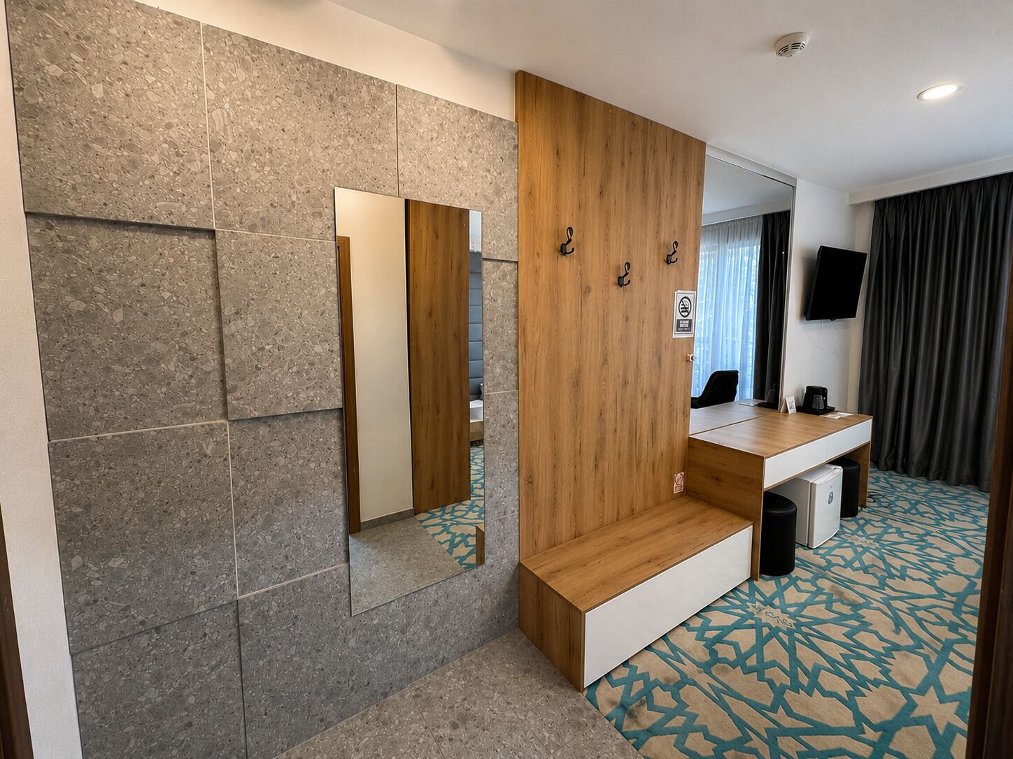 A modern hotel room with a large mirror, wooden panel wall, a small desk with a chair, a television, and a patterned carpet.