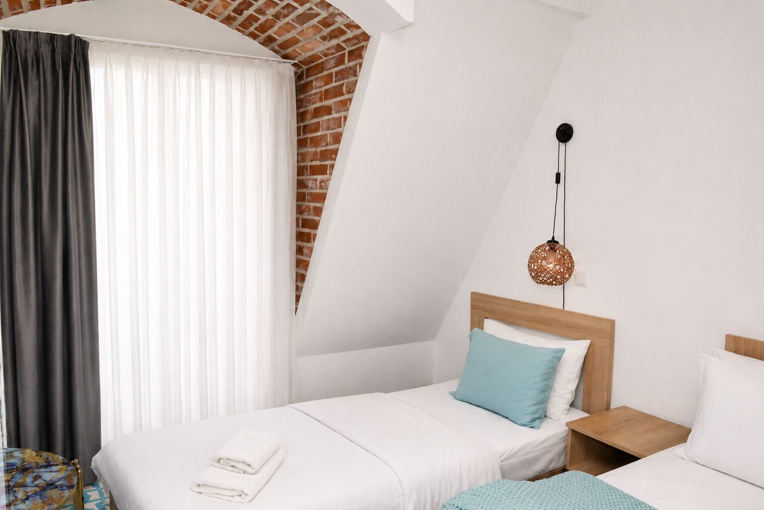 A hotel room with a sloped ceiling, a bed with white bedding, a blue pillow, a wooden nightstand, and a hanging light fixture.