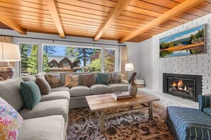 A cozy living room with a sectional sofa, a fireplace, and large windows offering a view of a house and trees. - Zephyr Cove Gem - 2-min walk to HOA Beach! (Zephyr Cove)