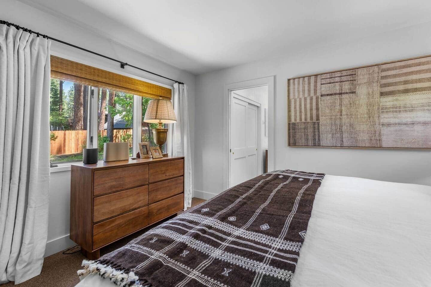 A bedroom with a wooden dresser, a bed with a patterned blanket, a window with curtains, and a wall-mounted artwork.