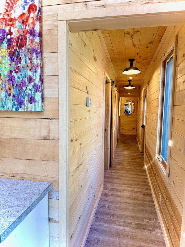 A wooden hallway with a painting on the left wall, a ceiling light, and windows on the right. - NEW!! Sandy Lakefront Cabin – Sleeps 10 with Kayaks & ATV Access (Fence)