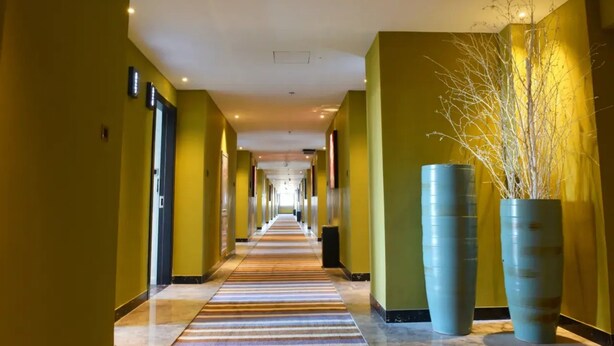 Un pasillo de hotel con alfombra a rayas, paredes amarillas y dos jarrones grandes a la derecha.