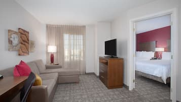 A hotel room with a sofa, a TV on a wooden stand, a bed with a red headboard, and a window with sheer curtains.