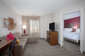 A hotel room with a sofa, a TV on a wooden stand, a bed with a red headboard, and a window with sheer curtains. - Suite w/ Full Kitchen | Near DEN | Free Breakfast (Denver)