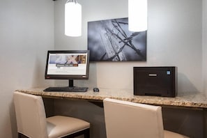 A hotel room with a desk, two chairs, a computer monitor, a desktop computer, and a framed picture on the wall.