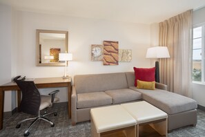 A hotel room with a desk, office chair, sofa, ottoman, and a large window with curtains. - King Suite Near Denver Airport | Free Breakfast (Denver)