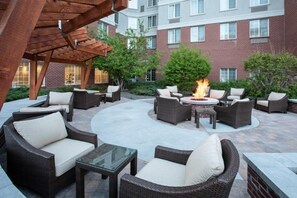A patio area with wicker furniture, a central fire pit, and a brick building in the background.