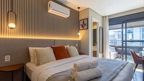 A hotel room with a bed, bedside tables, an air conditioning unit, a wall-mounted light, and a view of the city through large windows. - House in Condo. w/ Garage in Salvador Tstedf0401 (Salvador)