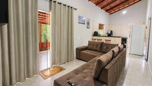 Una sala de estar con un sofá, un control remoto en el suelo, una zona de cocina con refrigerador y una mesa de comedor con sillas.