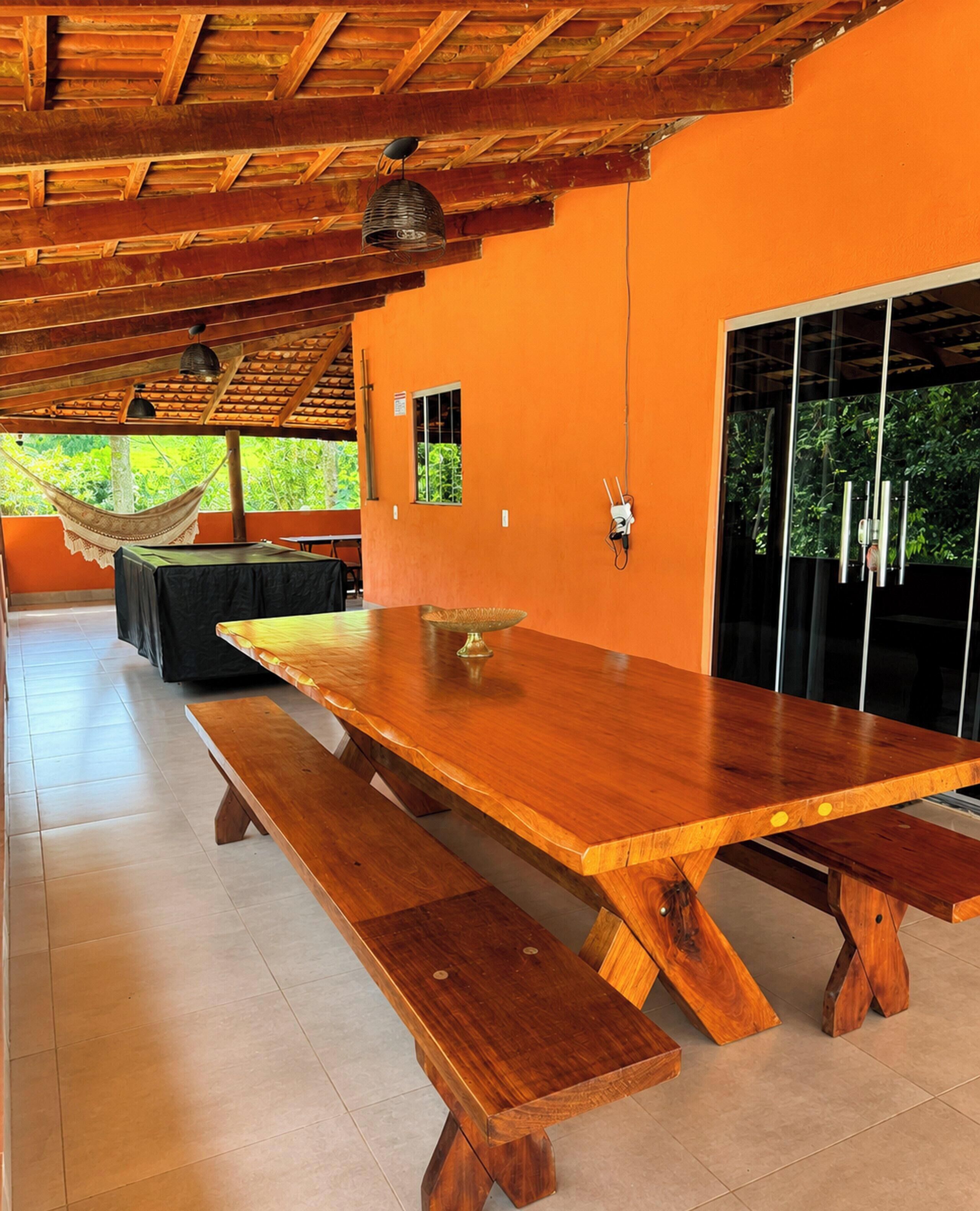 Uma mesa de jantar de madeira com bancos, uma rede e vista para o verde lá fora.