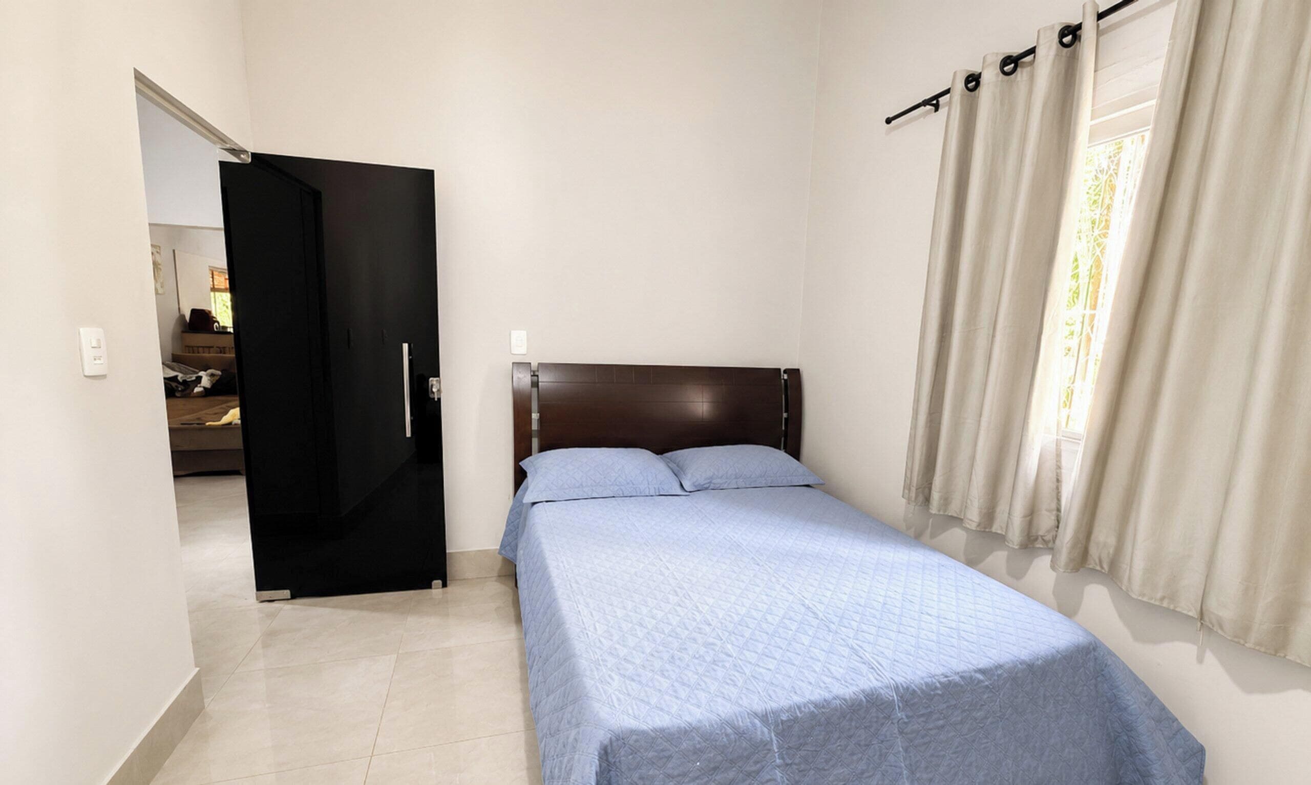 A bedroom with a bed, a door, a window with curtains, and a view into another room.
