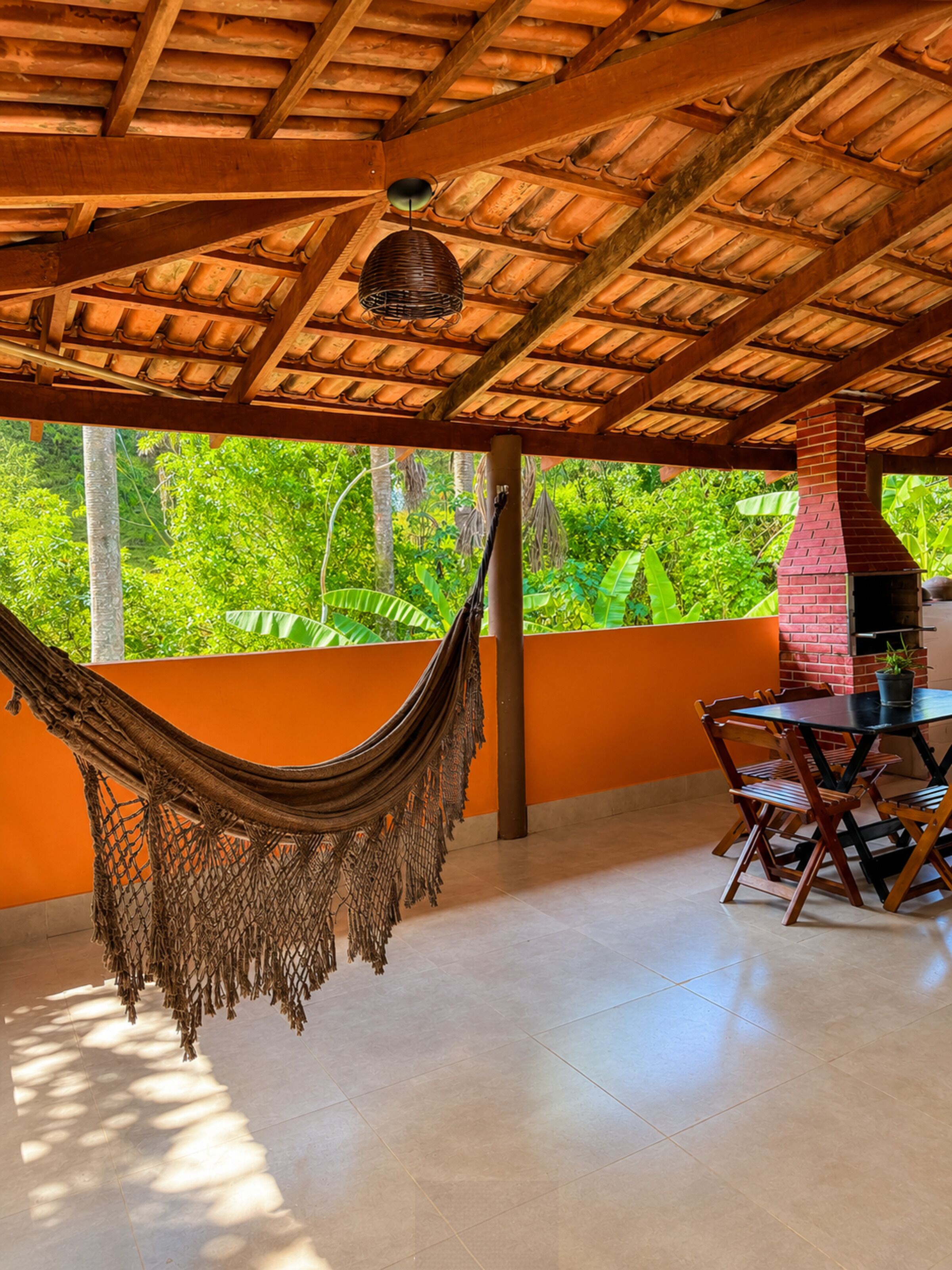A hammock, a brick chimney, and a table with chairs in a covered outdoor area with a view of greenery.