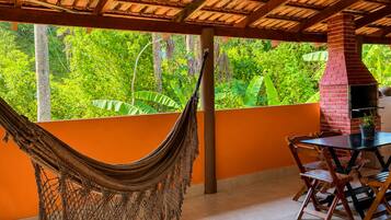 A hammock, a brick chimney, and a table with chairs in a covered outdoor area with a view of greenery.