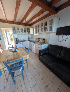 Une cuisine avec des poutres en bois, des meubles blancs, une table à manger avec des chaises, un canapé en cuir noir et un téléviseur fixé au mur.