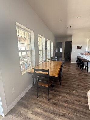 A spacious dining area with a long wooden table and chairs, large windows, and a kitchen area with bar stools.