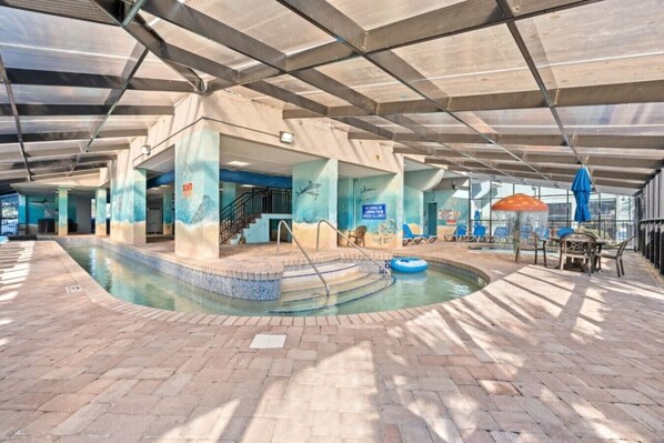 Una piscina interior con suelo de baldosas, un trampolín y una zona de estar cubierta con mesas y sillas.