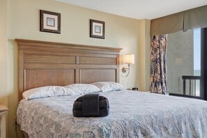 Ein Hotelzimmer mit einem großen Bett, zwei gerahmten Bildern an der Wand, einem Nachttisch, einer Lampe und Blick auf den Ozean durch ein Fenster mit Vorhängen.