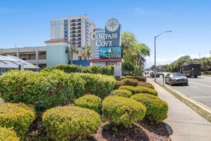 A hotel sign with a compass logo, a building with multiple floors, and a road with vehicles. - Compass Cove Resort 462 (Myrtle Beach)