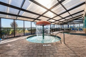 An indoor swimming pool with a water fountain, surrounded by a brick floor and glass walls offering a view of the outdoors.