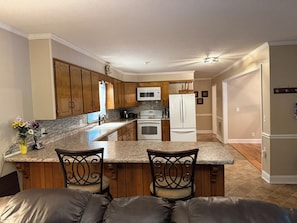A kitchen with wooden cabinets, a granite countertop, a white refrigerator, and a dining area with two chairs.