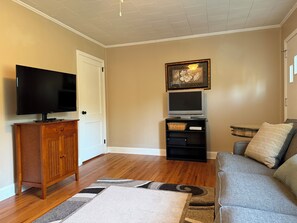 Ein Wohnzimmer mit Flachbildfernseher, einer Holzkommode, einem grauen Sofa, einem gemusterten Teppich und einem gerahmten Bild an der Wand.