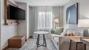 Una habitación de hotel con una televisión de pantalla plana, una mesa redonda, un sofá, un pequeño reposapiés, una lámpara y un cuadro en la pared.
