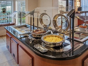 A buffet setup with various food items, including a bowl of scrambled eggs, a container of meat, and a bowl of fruit.