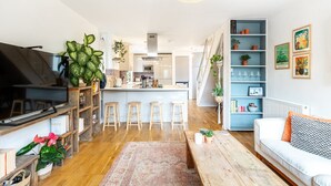 Un soggiorno moderno con un divano, un tavolino da caffè e sgabelli da bar. C'è un angolo cottura con fornello, forno a microonde e bancone. La stanza ha un pavimento in legno ed è decorata con piante e opere d'arte.