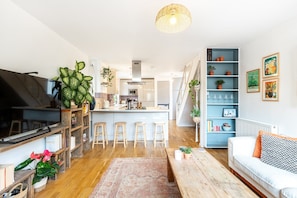A modern living room with a sofa, coffee table, and bar stools. There is a kitchen area with a stove, microwave, and bar counter. The room has wooden flooring and is decorated with plants and artwork. - Stylish Clapham Retreat Near the Common (London)
