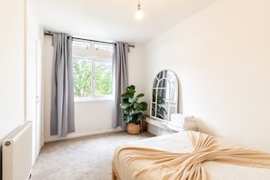 A bedroom with a bed, a window with curtains, a potted plant, a mirror, and a radiator. - Stylish Clapham Retreat Near the Common (London)