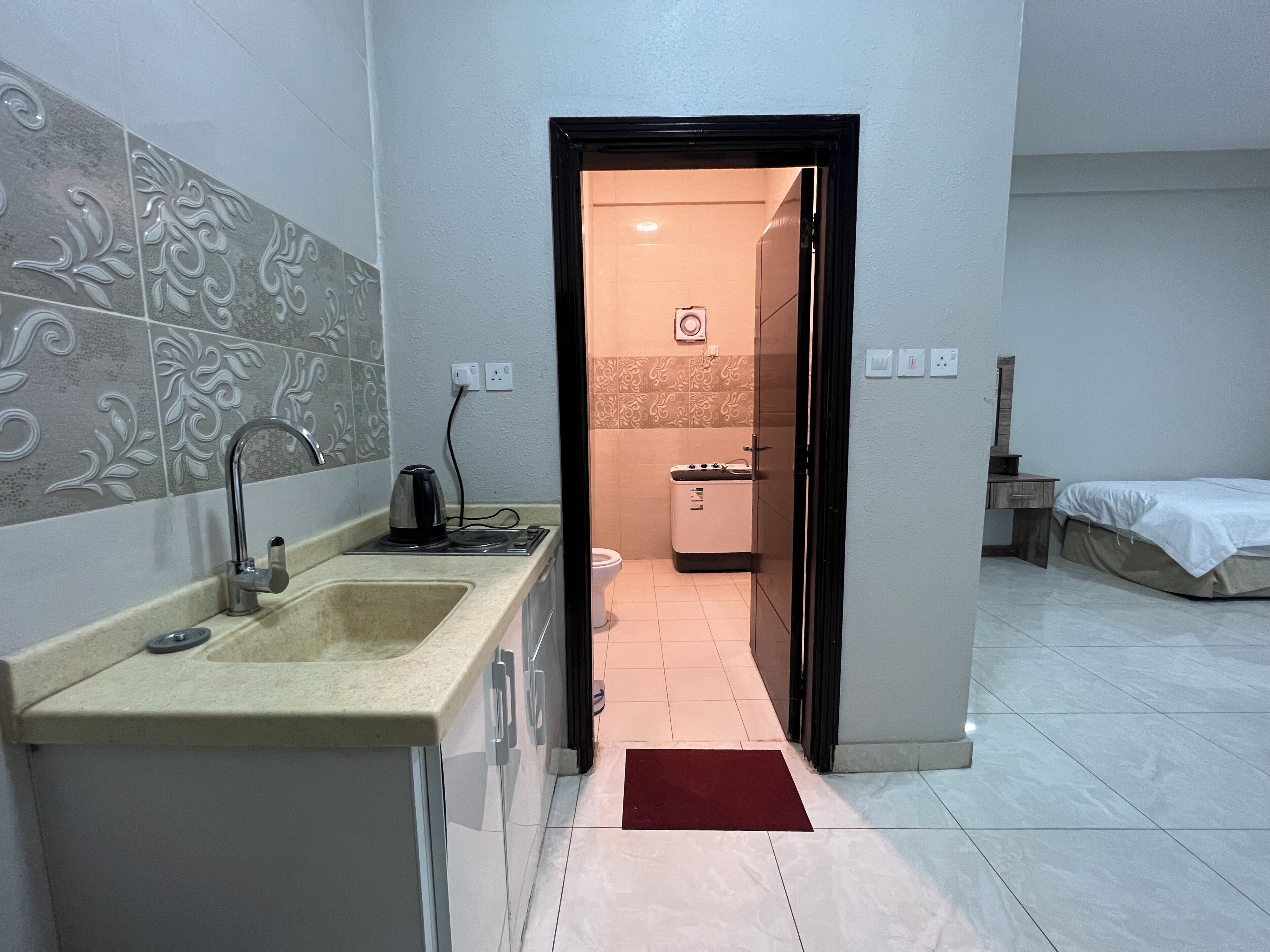 A modern kitchen with a sink, kettle, and stove, leading to a bathroom with a toilet and a shower.